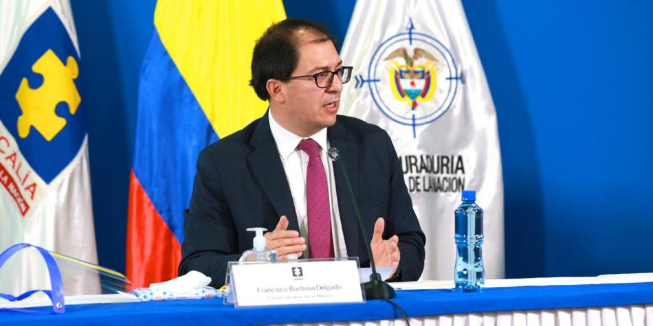 El fiscal general, Francisco Barbosa, sentado en una mesa, con las banderas del Colombia, Produradoría y Fiscalía de Fondo